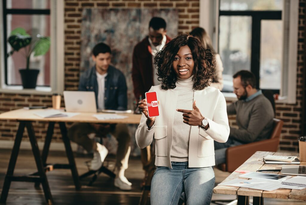 smiling african american casual businesswoman pointing at smartphone with youtube app in loft office